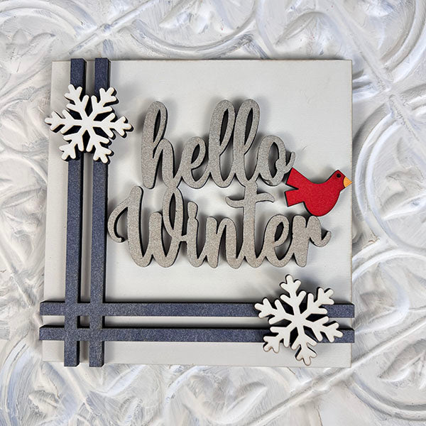 This winter-themed decorative sign from Janet's Craft Corner is perfect for enhancing your seasonal décor, whether used on tiered trays or as part of Winter Interchangeable Squares. It features the phrase "hello Winter" in gray script with a small red bird perched on the "t," and white snowflakes accentuating the corners against a textured white background.