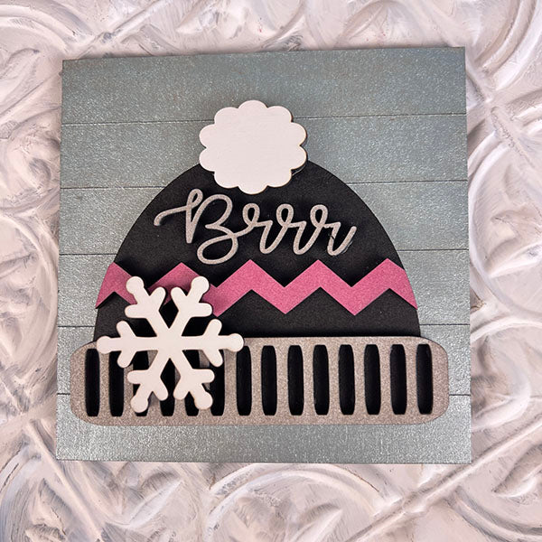 This decorative card from Janet's Craft Corner features a pink zigzag patterned winter hat with a pom-pom and the word "Brrr" written on it, set against a textured background. A white snowflake adorns the hat, making it an ideal addition to Winter Interchangeable Squares or tiered trays for your seasonal décor.