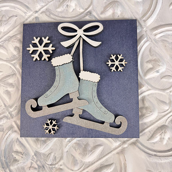 A decorative card showcasing a pair of silver ice skates tied with a bow, adorned with three white snowflakes. Ideal for seasonal décor, the background features Janet's Craft Corner's Winter Interchangeable Squares' iconic textured, swirling pattern.