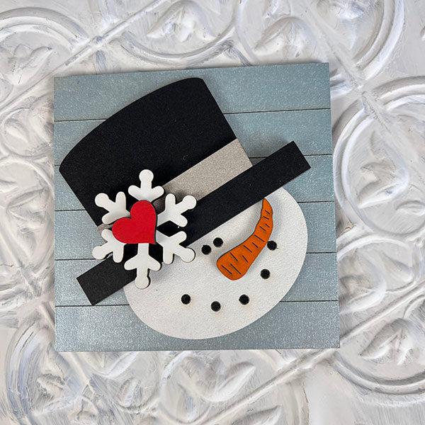 This decorative piece from Janet's Craft Corner's Winter Interchangeable Squares collection showcases a snowman face adorned with a black top hat, complete with a carrot nose and coal eyes and mouth. The design features a large white snowflake alongside a red heart on the hat, set against a textured light gray background, making it an ideal addition for tiered trays.