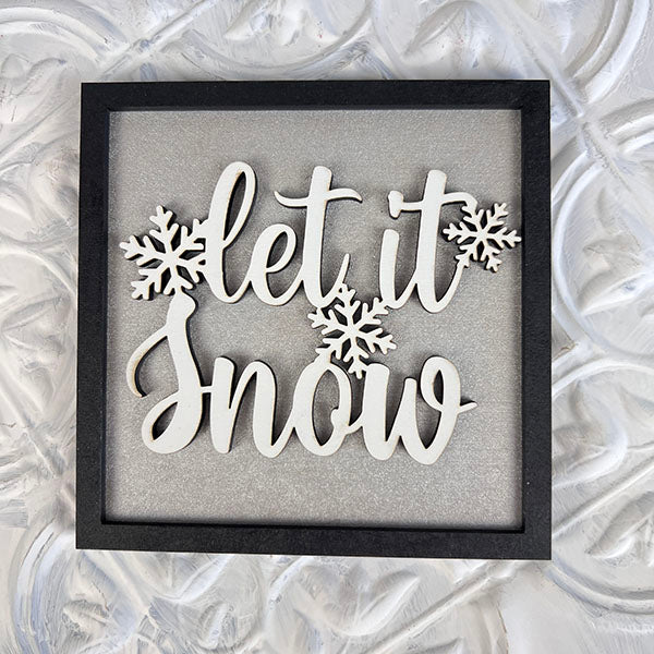 Introducing the Winter Interchangeable Squares by Janet's Craft Corner—a delightful framed piece featuring "let it snow" in elegant white cursive, complemented by charming snowflake designs set against a textured white backdrop. It's the perfect seasonal décor addition for your tiered trays.