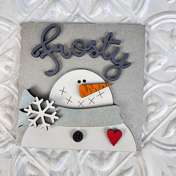 The "Winter Interchangeable Squares" by Janet's Craft Corner features a charming decorative piece that's ideal for tiered trays. This sign depicts a snowman with a carrot nose, buttons, and scarf, and is enhanced with a delicate snowflake and a red heart. The word "Frosty" is elegantly scripted above on a textured background, providing an enchanting touch to your seasonal décor.