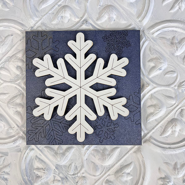 An elegant large white snowflake pattern on a textured dark background, encircled by smaller understated snowflakes, set against an embossed white backdrop. Ideal for enhancing your seasonal décor or showcasing in Janet's Craft Corner's Winter Interchangeable Squares on delightful tiered trays.