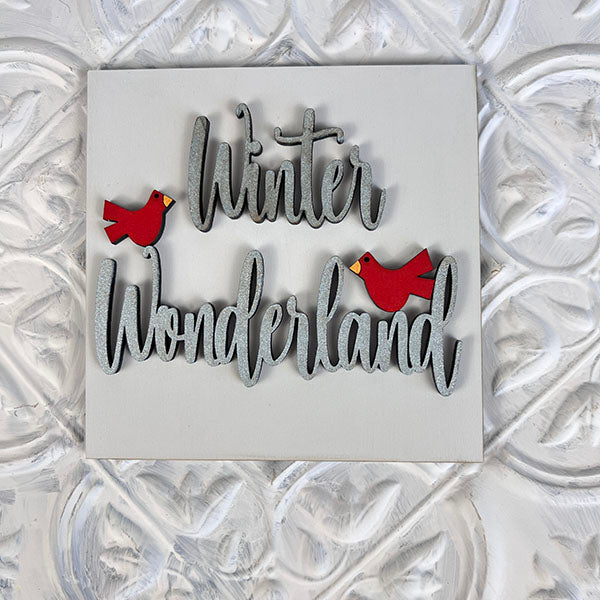 This decorative sign features the phrase "Winter Wonderland" in elegant gray script, ideal for seasonal décor with its charming illustrations of two small red birds, and mounted on a textured white wall. It makes an excellent addition to Janet's Craft Corner's Winter Interchangeable Squares or tiered trays for a festive touch.