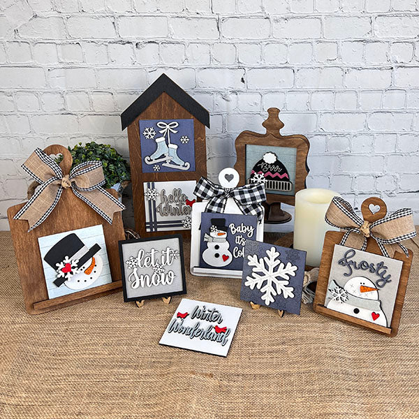 Introducing the Winter Interchangeable Squares from Janet's Craft Corner—a delightful assortment of seasonal décor. These winter-themed wooden decorations feature charming snowmen, delicate snowflakes, and festive phrases such as "Let it Snow" and "Hello Winter," all set against a brick wall backdrop. Perfect for enhancing your tiered trays, these pieces are designed with rustic finishes and adorned with bows to create a cozy, festive atmosphere.