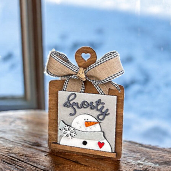 Add festive charm with Janet's Craft Corner Breadboard Swappable Squares Holder—DIY kit includes a small wooden sign featuring a snowman, "frosty" text, snowflake, and red heart. Perfect holiday décor for your table or snowy window display.