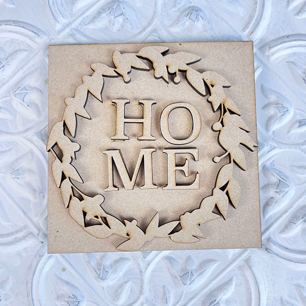 Introducing the "Home Family Interchangeable Squares" from Janet's Craft Corner. This delightful wooden square decoration showcases the word "HOME" intricately cut out in the center and surrounded by graceful leaf motifs. It sits beautifully on a textured, white surface, making it an ideal mini sign to enhance your home decor with warmth and style.