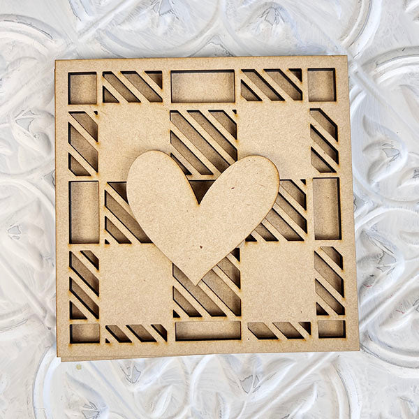 Introducing the Home Family Interchangeable Squares by Janet's Craft Corner—this delightful decorative wooden square boasts a central heart cutout and features an intricate pattern of mini squares and diagonal stripes. It's the perfect addition to your home decor, offering a touch of love on any textured white background.