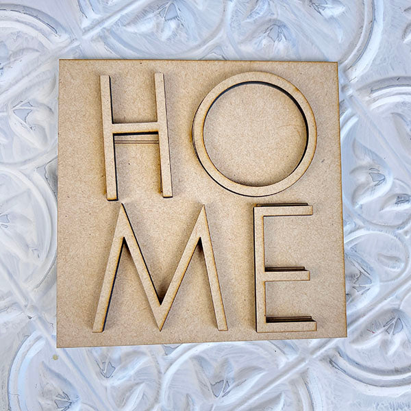 A wooden cutout spelling "HOME," part of the Home Family Interchangeable Squares collection from Janet's Craft Corner, sits on a textured, decorative white surface with swirling patterns. It adds a charming touch to your home decor and can be seamlessly incorporated into any room's aesthetic.