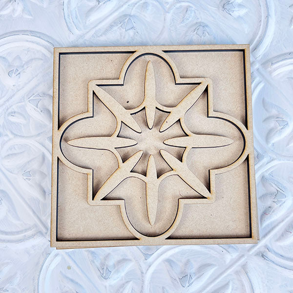 A square coaster from Janet's Craft Corner, branded as "Home Family Interchangeable Squares," is adorned with an intricate geometric star pattern. It rests on a textured white surface, adding elegance to the home decor. The design boasts symmetrical cut-out shapes, offering a decorative and artistic appearance that ideally complements interchangeable signs or mini sign displays.