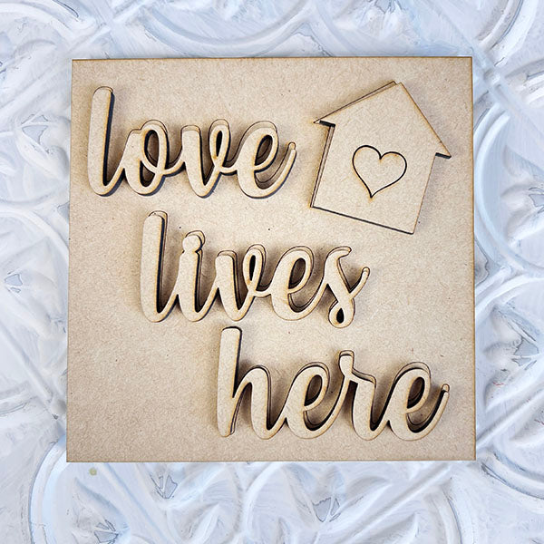 Introducing Janet's Craft Corner's "Home Family Interchangeable Squares" - a charming wooden plaque featuring cursive text that reads "love lives here." Designed in the shape of a small house with a heart cutout, it's perfect for mini signs that add character to any space. Set against a textured white background, this delightful piece makes an ideal addition to your home decor.