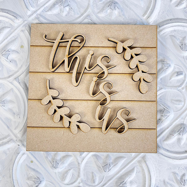 The "Home Family Interchangeable Squares" by Janet's Craft Corner is a charming wooden mini sign adorned with the phrase "this is us" in cursive, accented by decorative leaf designs. Its textured white surface showcases intricate patterns, making it an ideal addition to home decor with its versatile appeal.