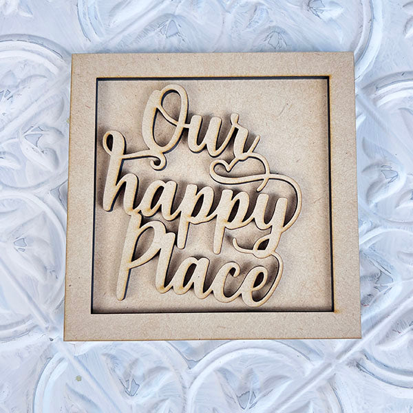 Introducing the "Home Family Interchangeable Squares" by Janet's Craft Corner—a delightful decorative wooden sign featuring a cursive font with the phrase "Our happy place." This charming mini sign adds warmth and style to any space as it rests elegantly on a textured white-gray surface, making it perfect for your home decor.