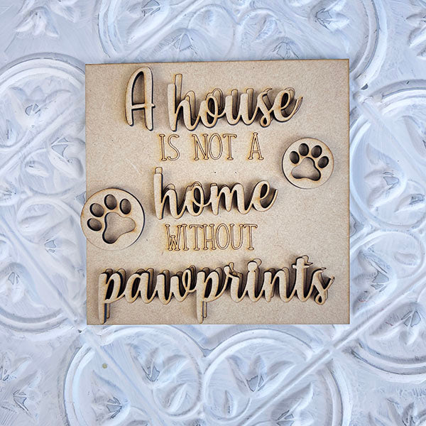 Introducing Janet's Craft Corner's Dog Interchangeable Squares: This charming mini wooden plaque features the heartwarming phrase, "A house is not a home without pawprints," adorned with two playful paw print designs. Perfect for farmhouse decor and dog enthusiasts, it stands out against a textured white background.