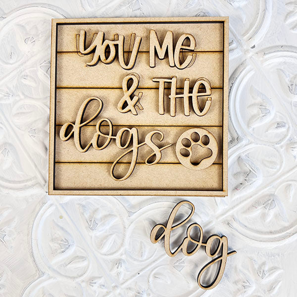 A wooden sign from Janet's Craft Corner, known as the "Dog Interchangeable Squares," gracefully displays the phrase "You Me & The Dogs" with a paw print on a light-patterned background. Ideal for enhancing tiered trays or infusing farmhouse charm into your decor, it features the word "dog" prominently in matching decorative style.