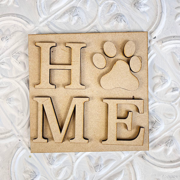 The "Dog Interchangeable Squares" by Janet's Craft Corner features a wooden sign with the letters "H," "M," and "E," alongside a paw print symbol replacing the "O" to spell out "HOME." This delightful piece, ideal for tiered trays, combines rustic farmhouse style with a tribute to our cherished pets, all set against a textured white background.