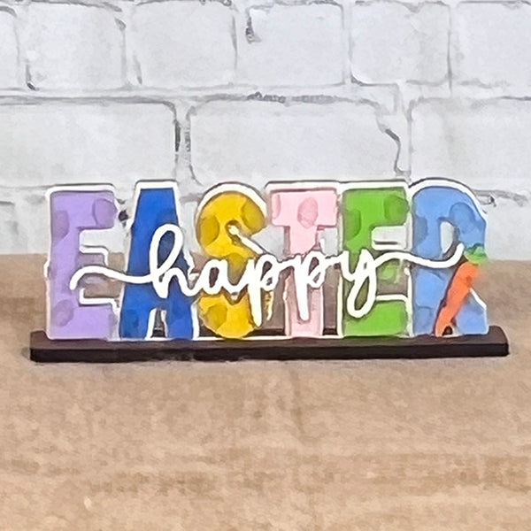 Introducing the Easter Trio Mini Shelf Sitters from Janet's Craft Corner: a delightful wooden Easter decoration featuring "EASTER" with colorful painted letters and a unique carrot design on the "R." The word "happy" beautifully scripted in cursive spans across the letters. This charming piece of decor is perfect for adding a festive touch to your shelf, complemented by its sturdy wooden base and set against a classic white brick background.