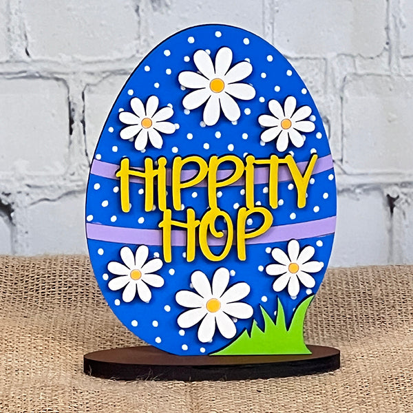 The "Easter Trio Mini Shelf Sitters" by Janet's Craft Corner features a charming blue egg-shaped sign adorned with white polka dots and daisy decorations, along with the playful "Hippity Hop" text in yellow. This delightful Easter decor piece pairs beautifully with a burlap surface and brick background, making it an ideal addition to your shelf sitters collection.