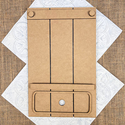 The Pallet Interchangeable Base, part of the DIY home decor kit by Janet's Craft Corner, features a blank brown surface with two clips and a circular hole at the bottom center. It is artfully positioned on a decorative, textured background, adding a charming touch to your home décor. The board is sectioned off with black lines on its wood or cardboard surface for added versatility.