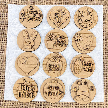 A set of twelve coasters from Janet's Craft Corner, featuring seasonal and themed engravings such as "Frozen Season," "Feb 14," "Trick or Treat," and "Give Thanks." The interchangeable designs include a bunny, heart, pineapple, and hat, all arranged in a 3x4 grid on a white background. This product is titled the Welcome to Our Home Door Hanger Interchangeable Set.