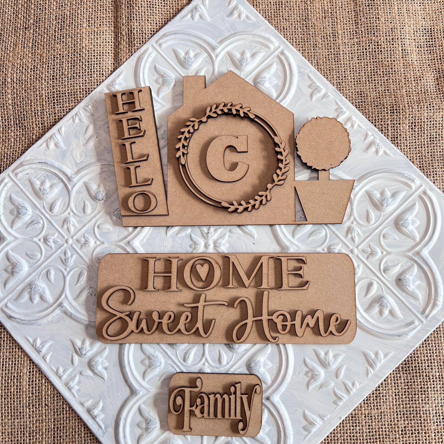 Crafted by Janet's Craft Corner, the Hello Home Monogram Interchangeable Insert | DIY home decor kit features wooden cutouts on a textured surface displaying a house with the word "Hello" and an interchangeable monogram insert, including a "C" wreath. Accompanied by accents like a potted tree and words such as "Home Sweet Home" and "Family," this scene provides a hand-painted DIY feel against the patterned fabric backdrop.