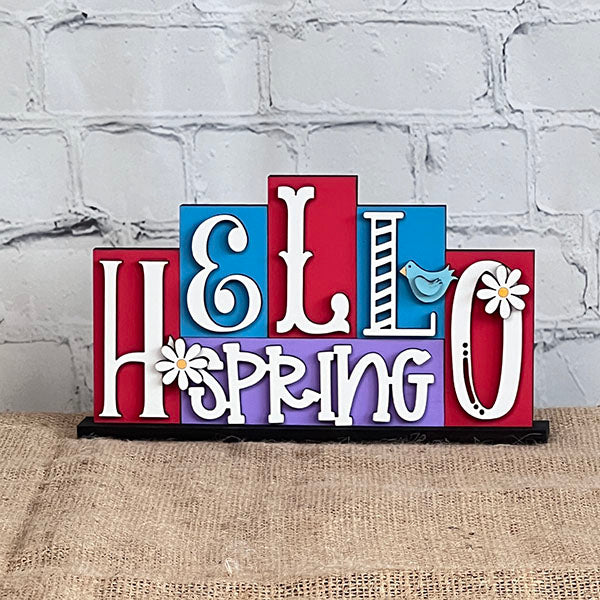 The Hello Spring Mini Word Block from Janet's Craft Corner features vibrant wooden lettering, with playful font variations. Ideal for home décor, it showcases a charming blue bird perched on the letter "R," and delightful daisies embellishing the "H" and "O." Positioned against a white brick wall on a burlap surface, it's perfect for adding a touch of spring to any room.