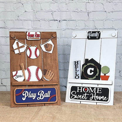 Displayed against a brick wall, two wooden decorative signs from Janet's Craft Corner's Pallet Interchangeable Base DIY home decor kit elegantly enhance your home décor. One sign features a baseball theme with "Play Ball" and "Home Run," while the other showcases a house motif with "Home Sweet Home," "Family," and a black house silhouette adorned with a green wreath.