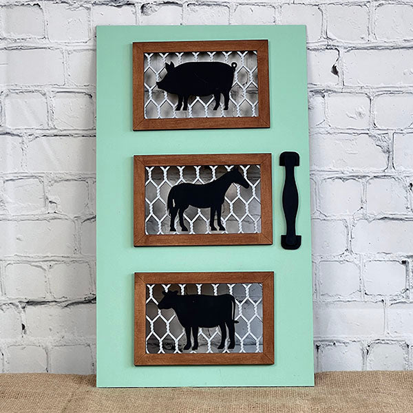 Introducing the Farm Animals Screen Door Sign by Janet's Craft Corner, a delightful decorative piece featuring three framed silhouettes of a pig, donkey, and cow set against a pastel green background with a textured white brick wall. Ideal for farmhouse decor, this wooden panel is accented with a black handle on the right side for added charm.