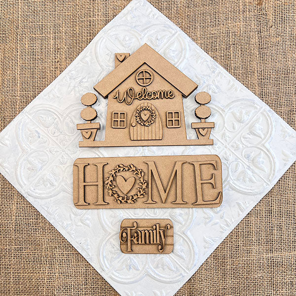 The "Welcome Home Interchangeable Insert" from Janet's Craft Corner is a DIY home decor kit that includes wooden cutouts of a house with signs reading "Welcome," "Home," and "Family." This charming piece features hand-painted details on a white textured square overlaying burlap fabric, with a heart wreath accenting the "Home" sign.