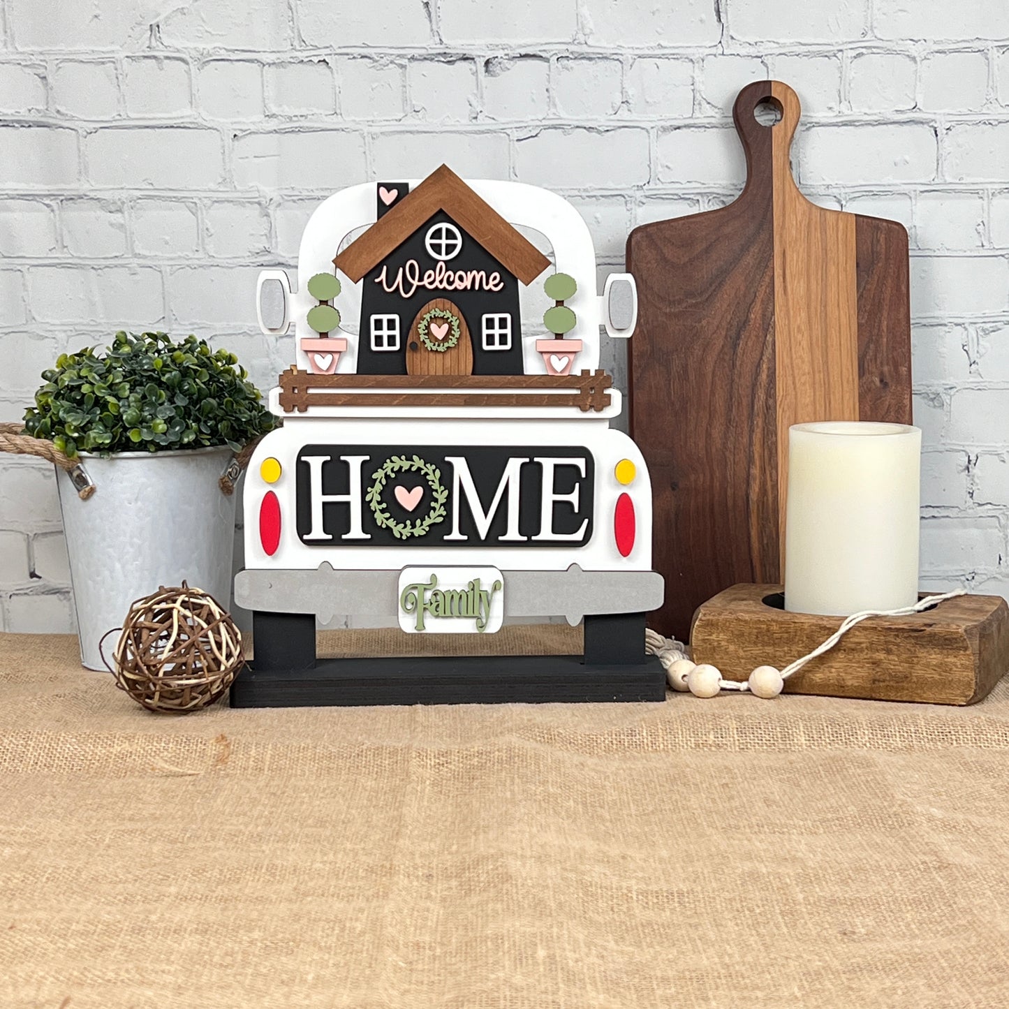 A decorative setup from Janet's Craft Corner showcases the DIY home decor kit, "Welcome Home Interchangeable Insert," featuring a hand-painted wooden truck sign highlighting the words "Welcome" and "Home." The display is enhanced with a potted plant, a wooden cutting board, a candle on a stand, and a woven ball against the backdrop of a brick wall. This customizable touch adds unique charm to any space.