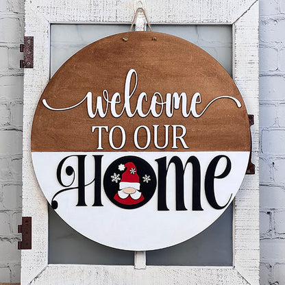 Introducing the Welcome to Our Home Door Hanger Interchangeable Set by Janet's Craft Corner. This decorative round sign enhances your home décor with an elegant "Welcome to our Home" design in white and black letters. The letter "O" in "Home" features a charming gnome wearing a red hat, adorned with snowflakes, perfect for adding a whimsical touch to your space. It hangs beautifully on any whitewashed wooden door.
