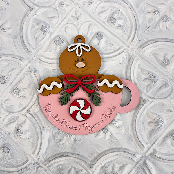 A gingerbread figure embellishes the delightful Gingerbread Teacup Ornaments - Painted, created by Janet's Craft Corner. This festive decoration showcases a peppermint candy design and features decorative text that reads "Gingerbread Kisses & Peppermint Wishes," set against a textured white background.