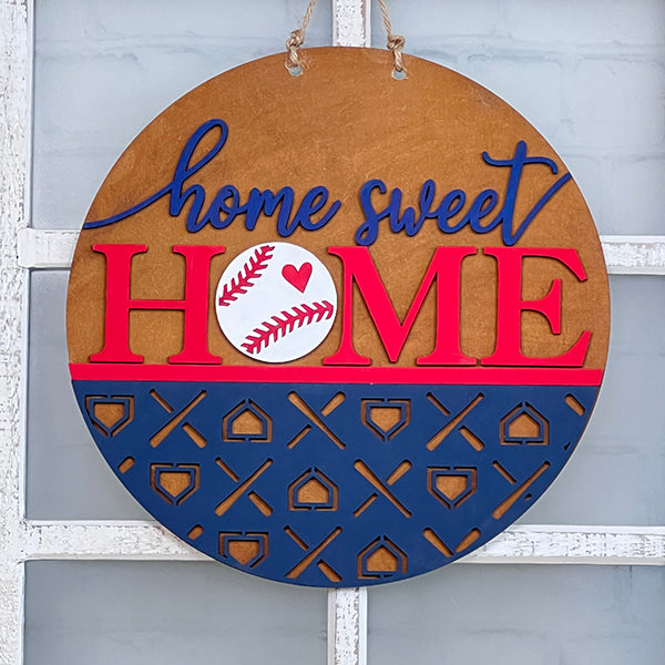 Crafted by Janet's Craft Corner, the Baseball Home Sweet Home Round is a round wooden door hanger featuring "home sweet HOME" emblazoned in blue and red. The "O" in "HOME" is cleverly designed as a baseball with a red heart, accented by a striking blue pattern. This delightful piece of home decor hangs elegantly by rope against a glass window and is an ideal addition for any DIY kit enthusiast.