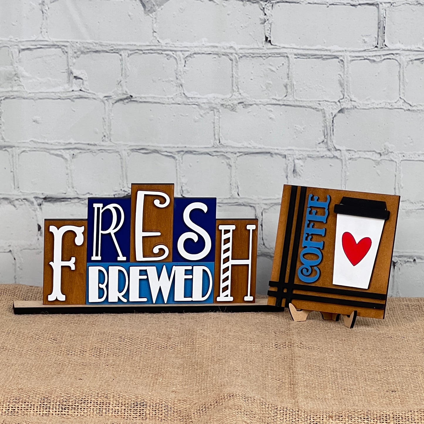 Janet's Craft Corner's Coffee Squares feature charming wooden signs on burlap with "FRESH BREWED" and "COFFEE" (cup and red heart) designs, perfect for DIY seasonal home decor in small spaces against a white brick wall.