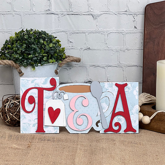 The Heartland Tea Word Block Shelf Sitter - Painted spells out “Tea” featuring a teacup, spoon, and heart design. This charming accent sits beautifully on a shelf or table decorated with potted plants, candles, and rustic touches against a white brick wall—perfect for tea enthusiasts who love cozy style.
