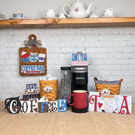 A charming coffee and tea station featuring a Keurig, mugs, baskets, teapots, and Janet's Craft Corner Tea Word Block Shelf Sitter DIY kit on a wooden shelf against a white brick wall—perfect for tea lovers.