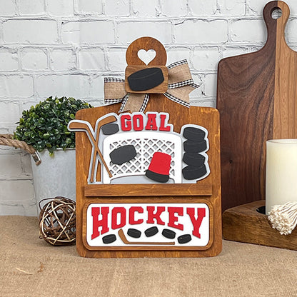 The Hockey Interchangeable Insert - Painted by Janet's Craft Corner is a decorative wooden sign featuring hockey-themed designs complete with a net, pucks, and sticks. The bold red words "Goal" and "Hockey" stand out on a white brick background, accented with a ribbon. Paired with a candle and wooden boards, this piece is ideal for adding charm to your home decor.