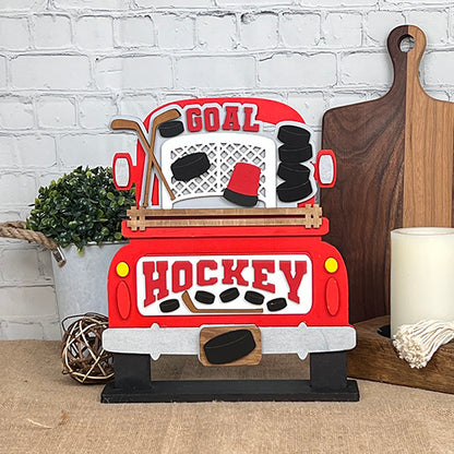 A delightful home decor piece from Janet's Craft Corner, the "Hockey Interchangeable Insert - Painted" showcases a red truck carrying "Goal Hockey" on its back. The truck bed is filled with hockey sticks, pucks, and a net, set against a brick wall backdrop. This charming insert is perfect for enhancing your space alongside plants and cutting boards.