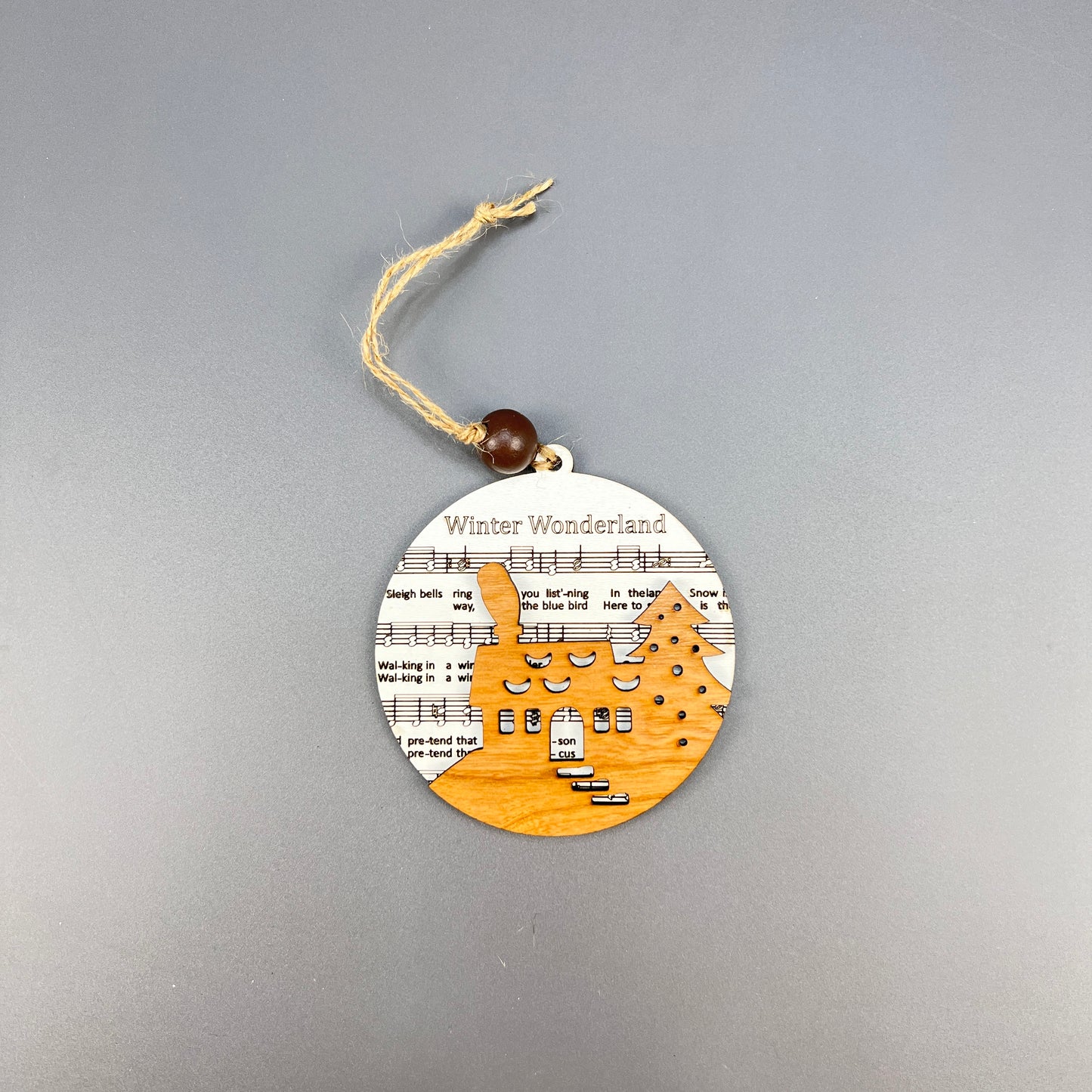 Discover the rustic charm of Janet's Craft Corner's Sheet Music Ornament, ideal for music enthusiasts. This ornament showcases a laser-cut wooden reindeer amidst snow-covered trees, all set against the backdrop of sheet music titled "Winter Wonderland." A twine loop with a wooden bead finishes off this charming piece.