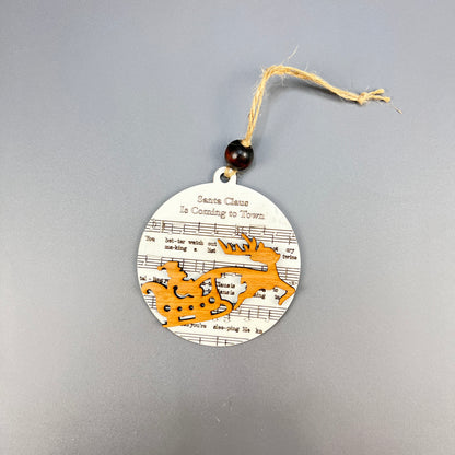 Immerse yourself in the rustic charm of Janet's Craft Corner with their Sheet Music Ornaments, featuring a round design that beautifully showcases a wooden cutout of Santa in a sleigh pulled by reindeer. Perfect for music lovers, the background is adorned with sheet music displaying "Santa Claus is Coming to Town." A convenient string at the top allows for easy hanging.
