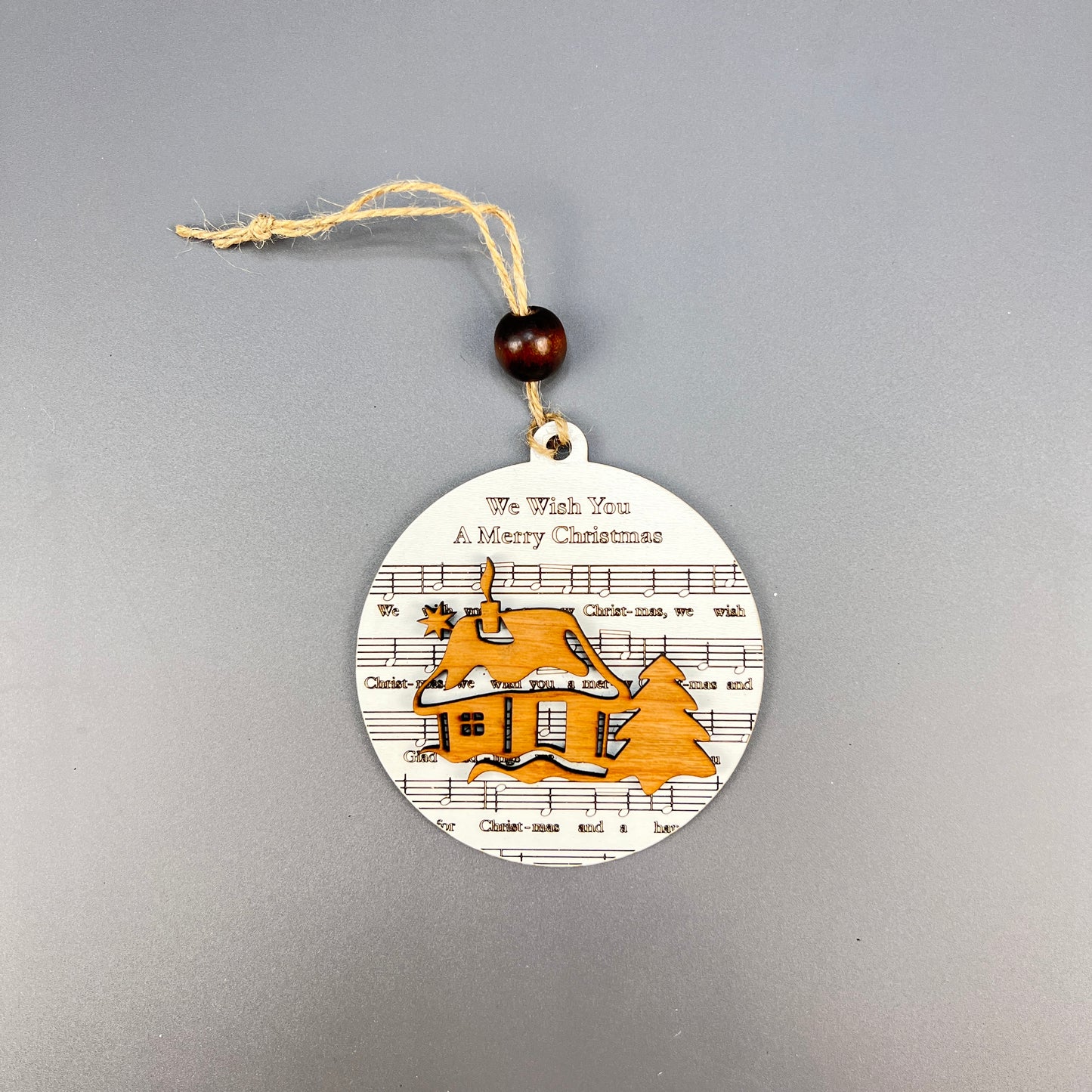 The Sheet Music Ornaments from Janet's Craft Corner exude rustic charm with a string hanger, showcasing sheet music along with the phrase "We Wish You A Merry Christmas." The ornament depicts a cottage surrounded by trees and snow in warm orange tones that music lovers will cherish.