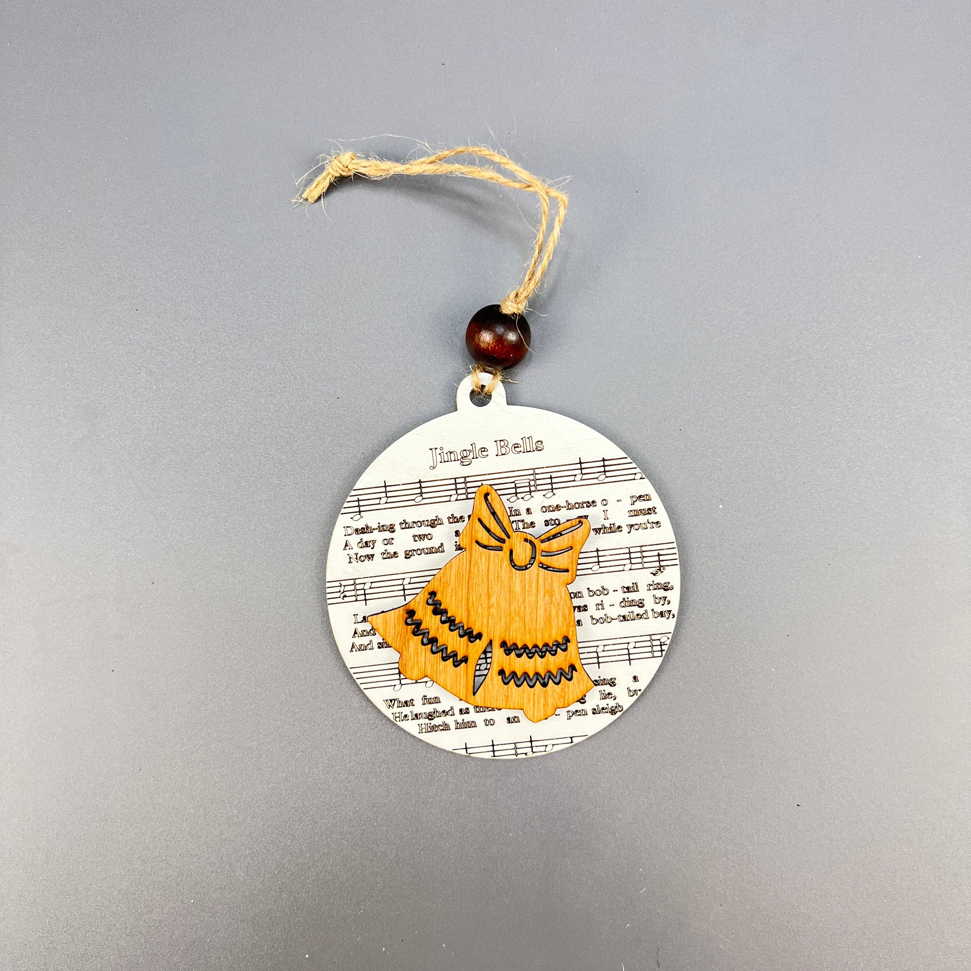 Introducing the Sheet Music Ornaments by Janet's Craft Corner: a delightful round ornament ideal for music enthusiasts, showcasing "Jingle Bells" on sheet music and a charming illustration of a yellow bell with a bow. Its rustic appeal is enhanced by a jute string and wooden bead for easy hanging.