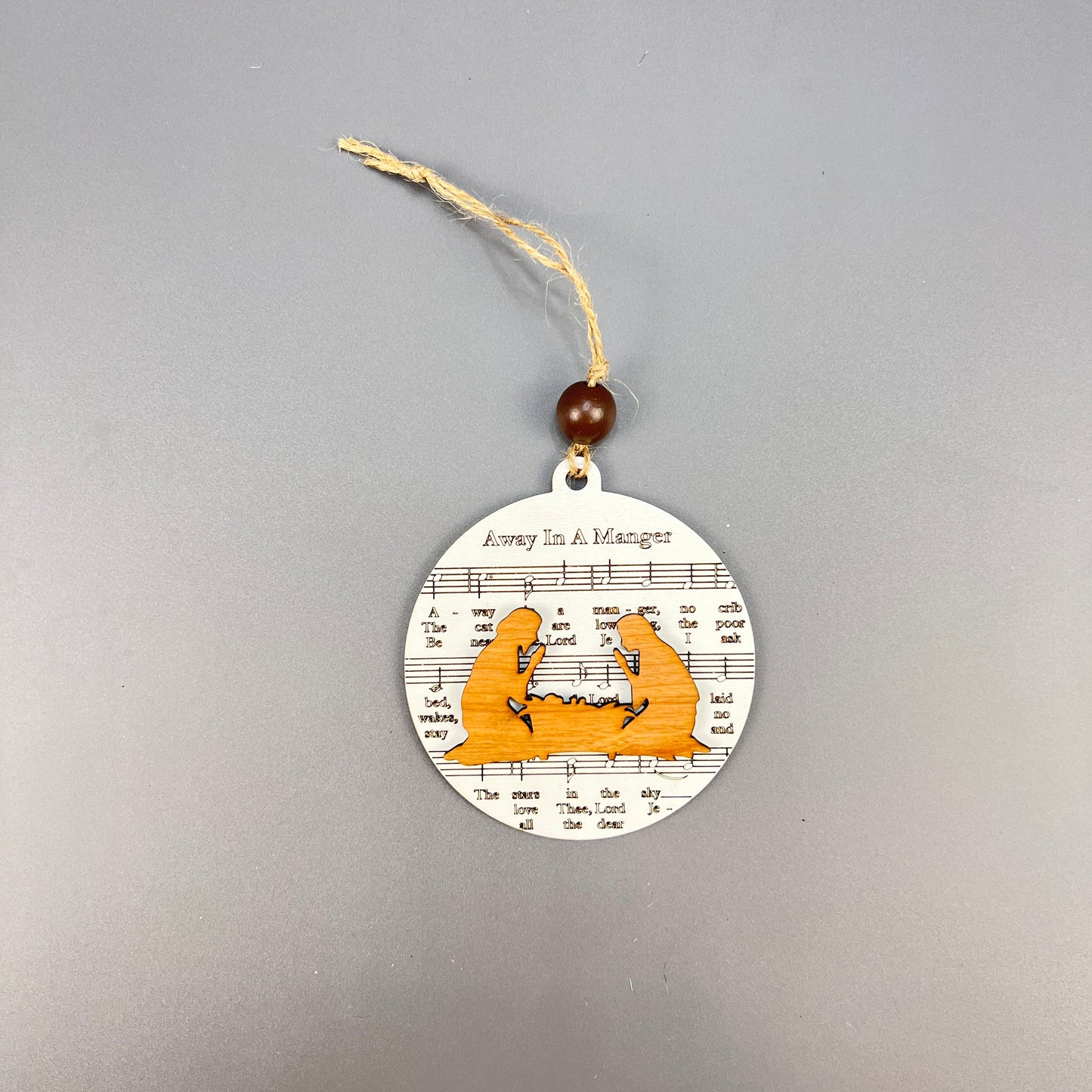 The Sheet Music Ornaments by Janet's Craft Corner offer rustic charm with a round design featuring a nativity scene, complete with a manger and baby depicted in an orange silhouette. The ornament's background showcases the phrase "Away In A Manger" at the top, making it ideal for music lovers. It comes with a string and wooden bead for easy hanging.