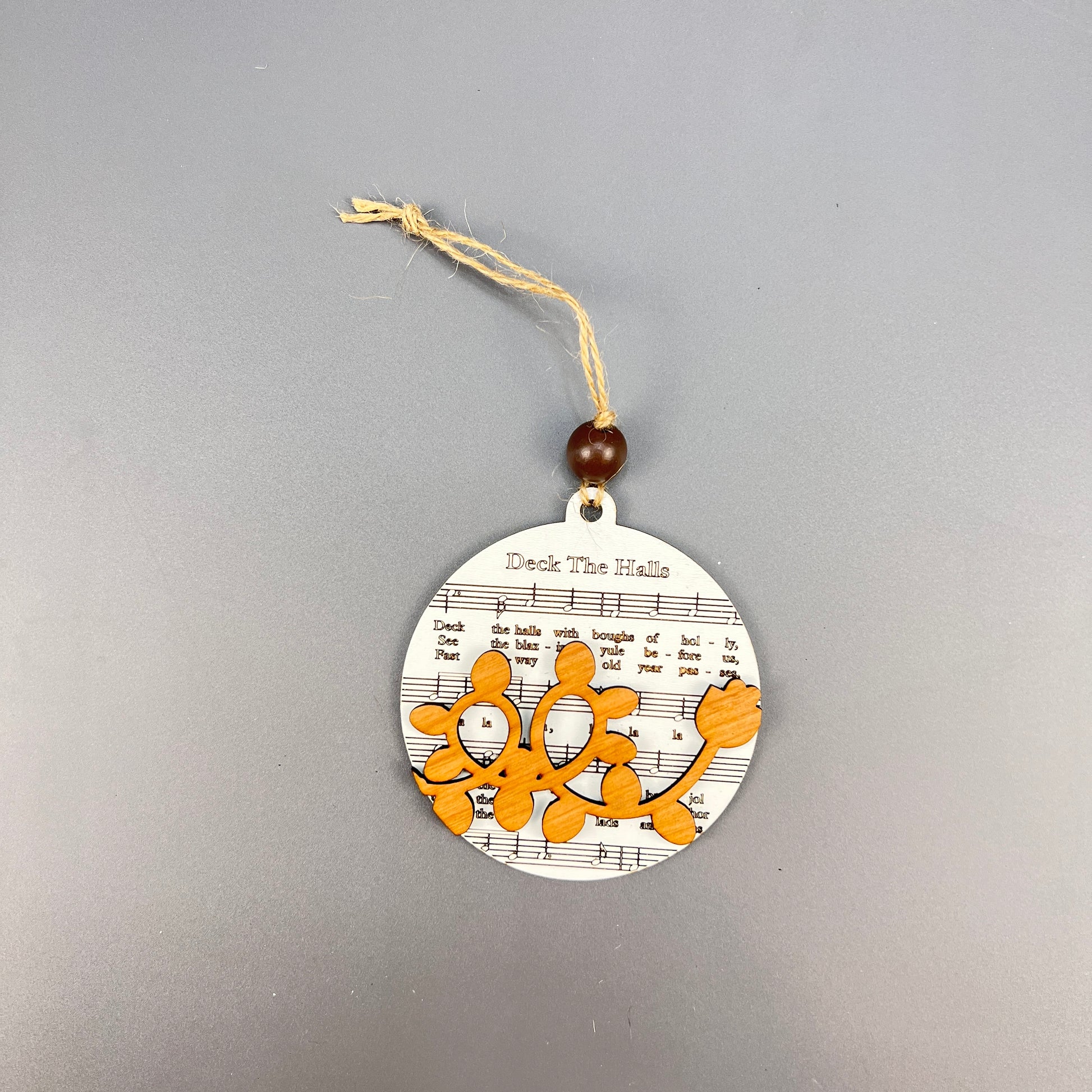 Janet's Craft Corner's Sheet Music Ornaments feature a circular design with a wooden gingerbread figure set against "Deck the Halls" sheet music, making it an ideal choice for music lovers. This ornament adds rustic charm with its twine and bead loop, perfect for hanging.