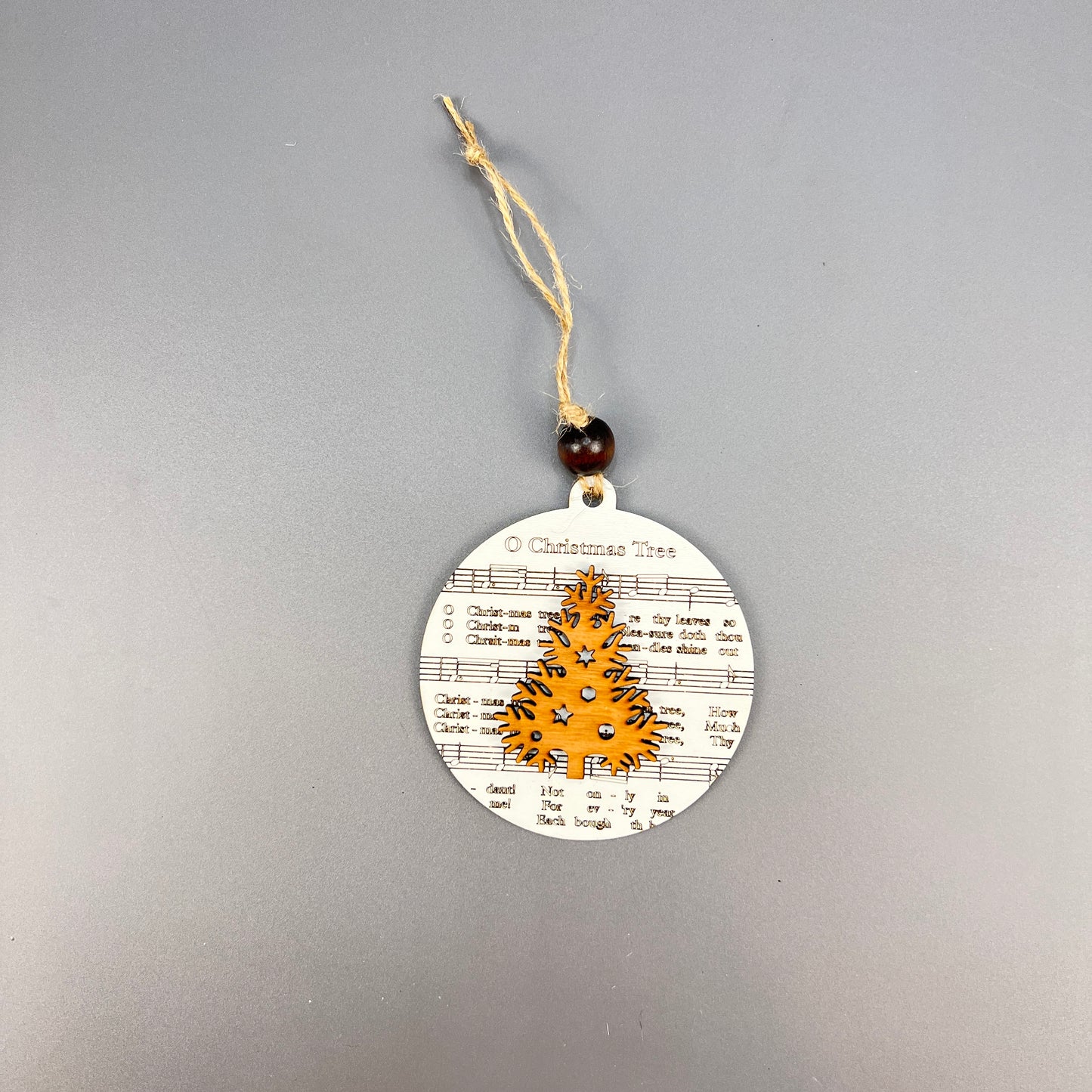 The Sheet Music Ornaments from Janet's Craft Corner are round Christmas decorations beautifully adorned with musical notes and lyrics, making them ideal for music enthusiasts. They showcase a golden tree at the center and exude rustic charm by hanging from a string with a wooden bead on a gray background.