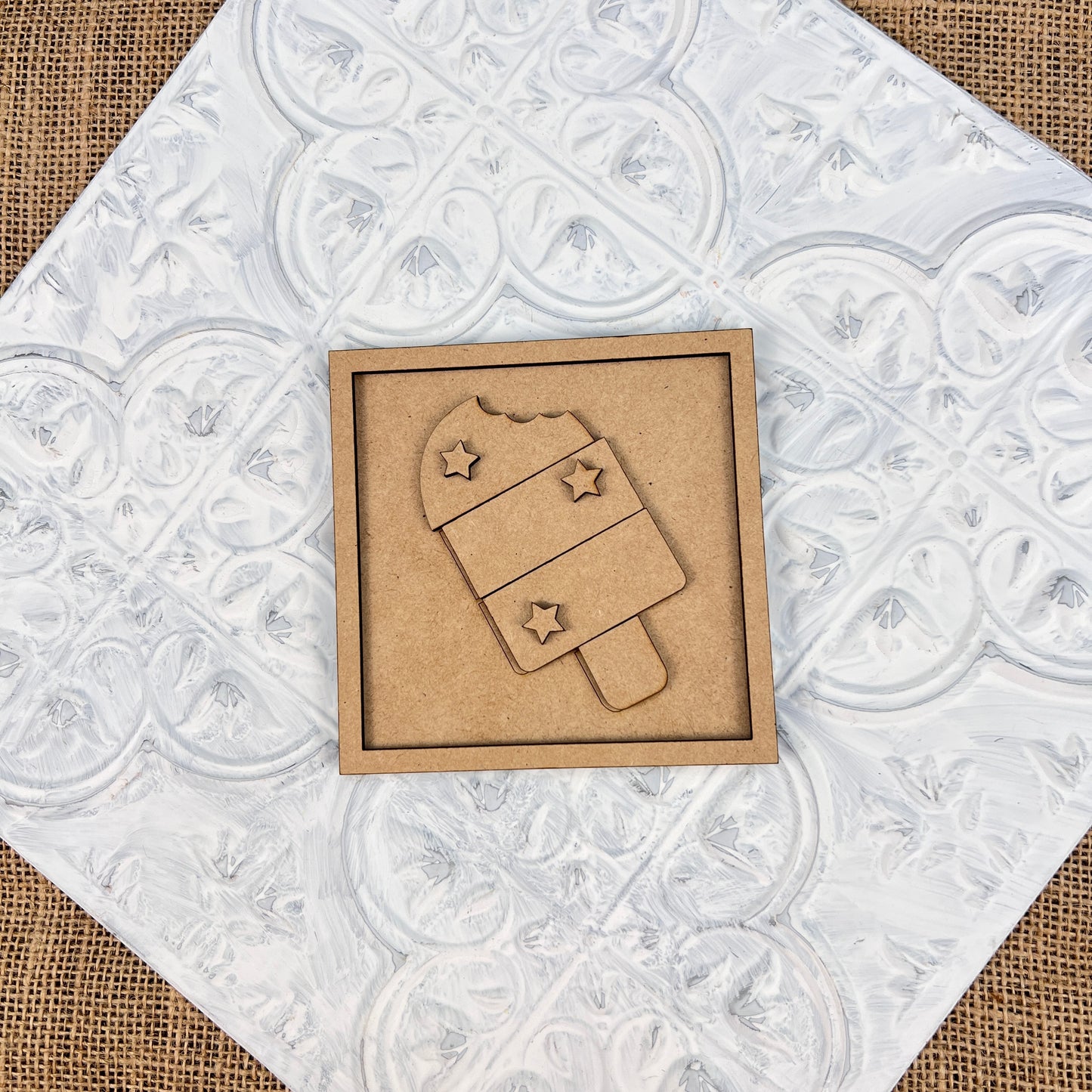 A laser-cut wooden piece featuring the Patriotic Interchangeable Squares design from Janet's Craft Corner sits on a textured white square tile. The design showcases a patriotically themed popsicle with stars and stripes, complete with a bite mark at the top. It is set against a charming burlap surface, perfect for farmhouse décor.