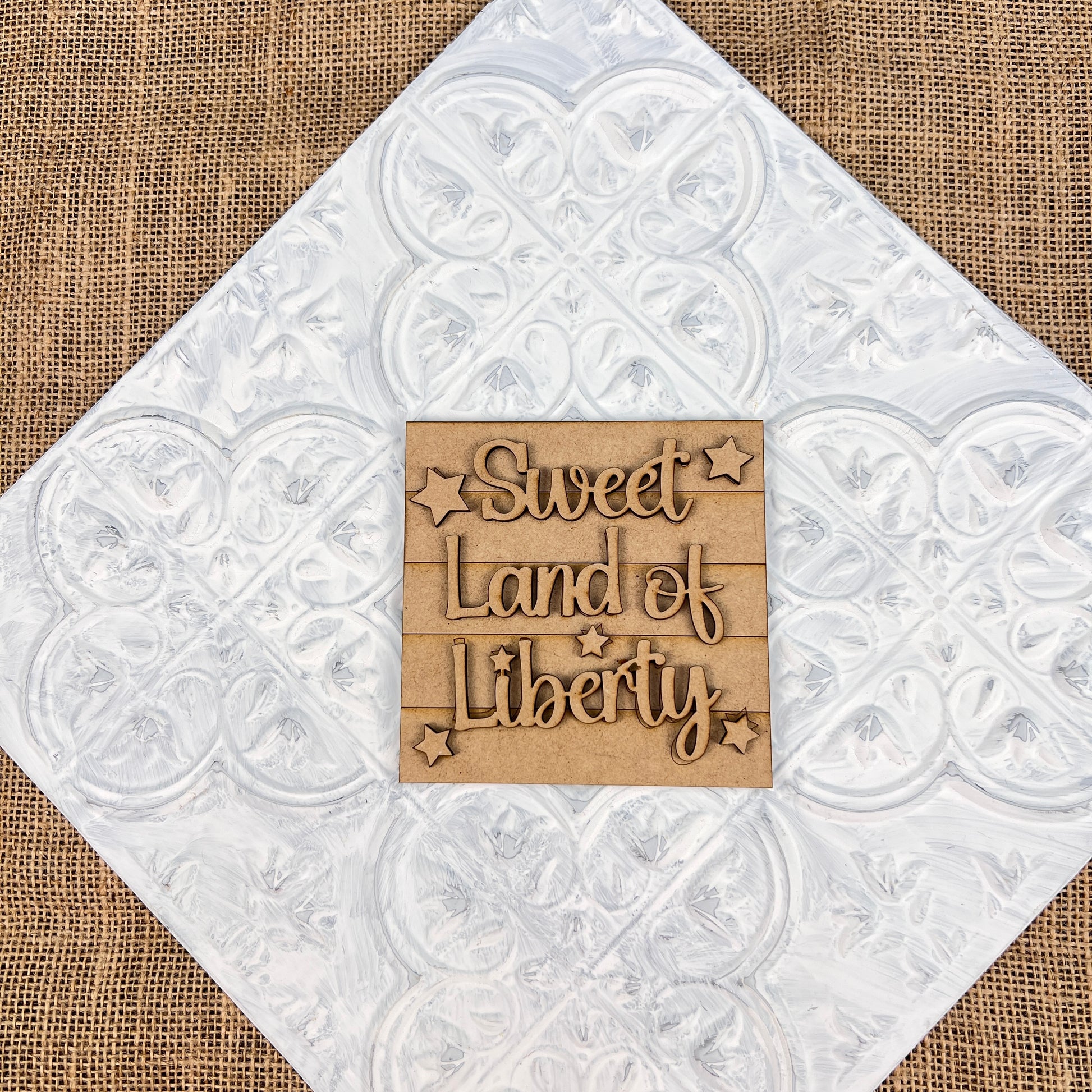 The "Patriotic Interchangeable Squares" by Janet's Craft Corner features a decorative piece with "Sweet Land of Liberty" carved in wood and adorned with small stars, set against a textured white square surface. The burlap material background enhances the farmhouse décor with a touch of seasonal flair.