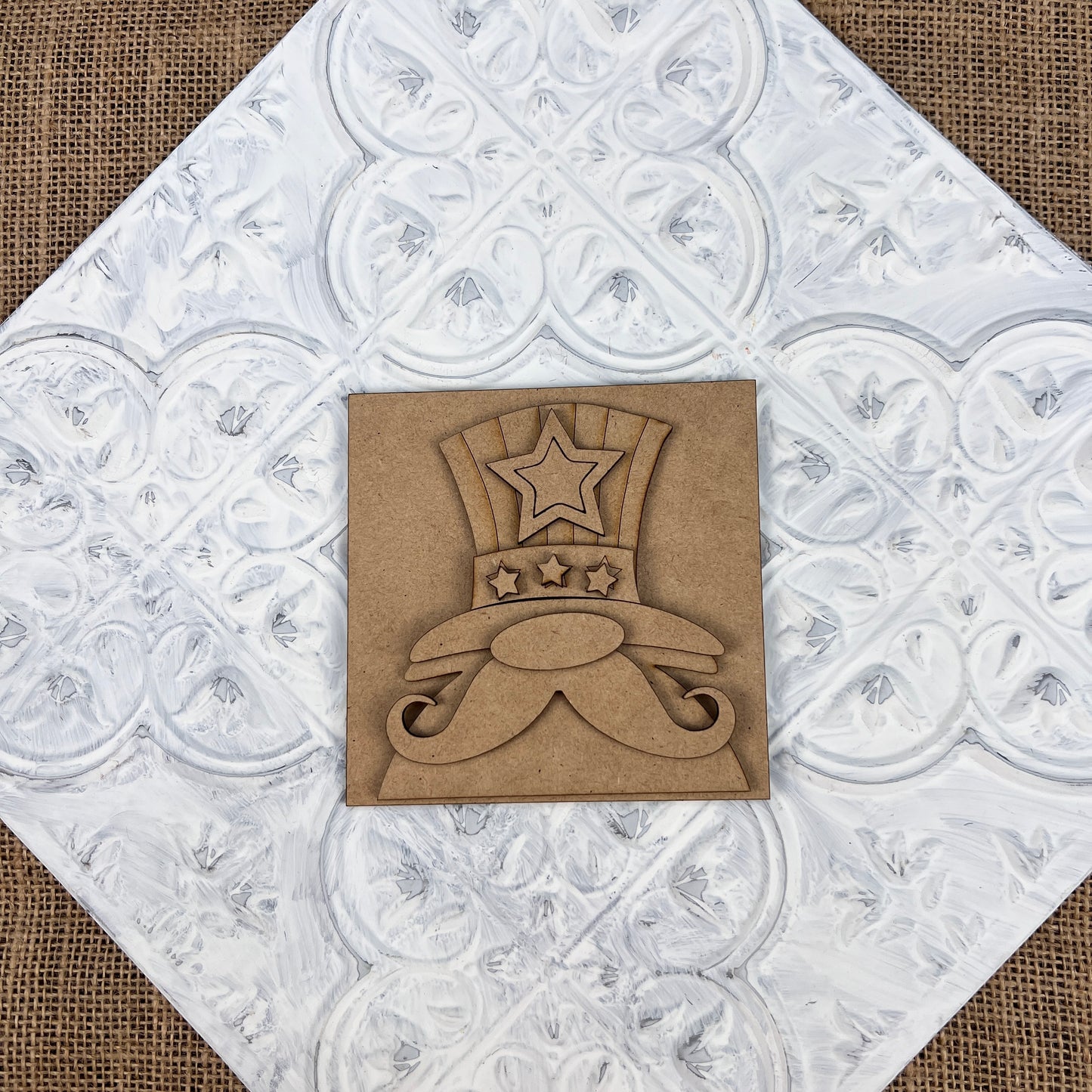 The Patriotic Interchangeable Squares by Janet's Craft Corner is ideal for farmhouse décor, featuring a decorative design with a bold top hat and mustache embellished with stars. It is set against a white textured background with detailed swirling patterns, adding a festive touch, and the plaque is positioned diagonally.