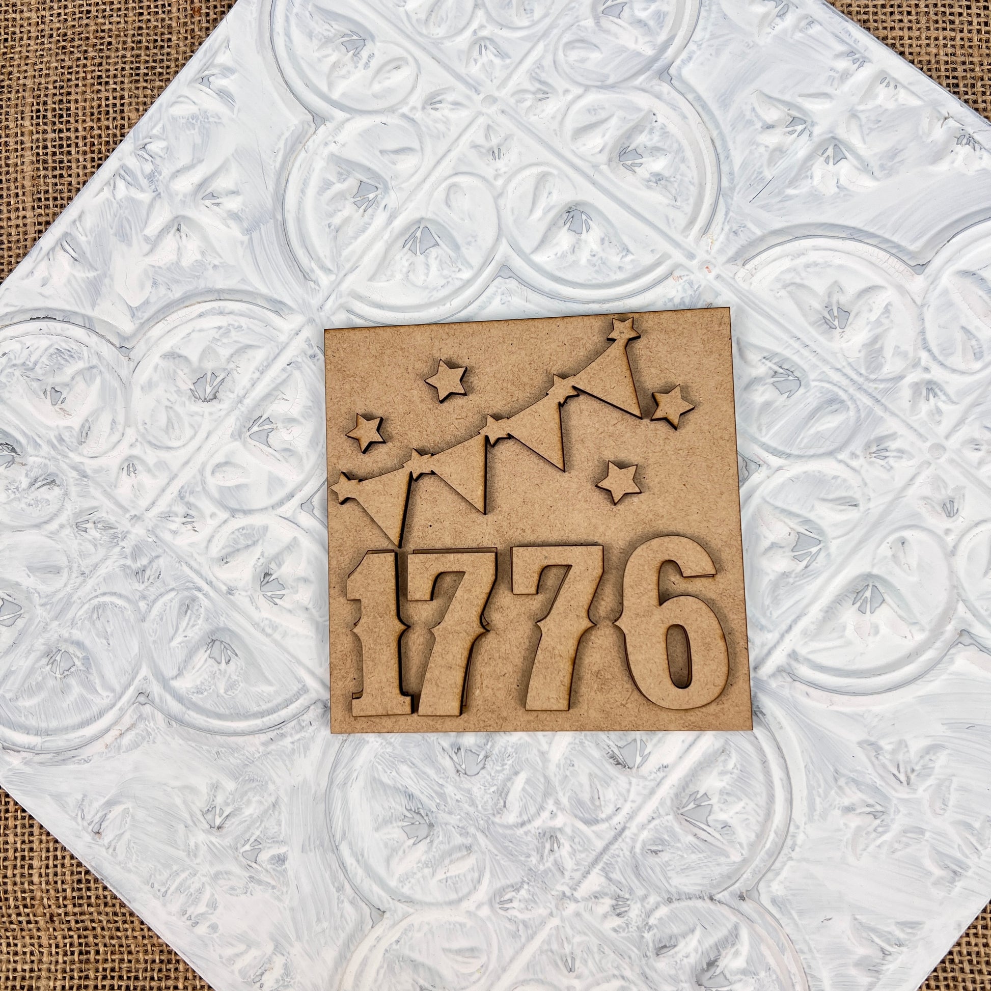 The "Patriotic Interchangeable Squares" from Janet's Craft Corner, an exquisite wooden ornament ideal for farmhouse décor, features the numbers "1776" with star and garland cutouts. This delightful design adds seasonal charm as it gracefully rests on a white, embossed surface.