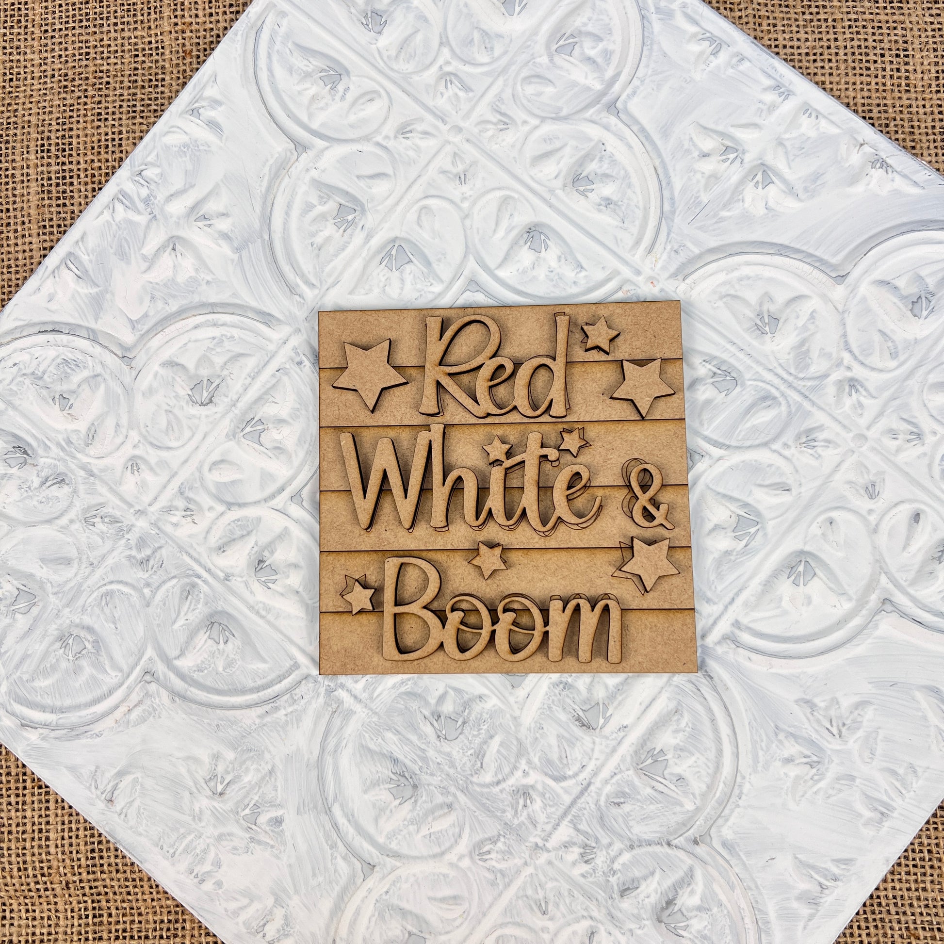 Wooden sign from Janet's Craft Corner's "Patriotic Interchangeable Squares" collection, featuring the phrase "Red White & Boom" surrounded by stars. The background boasts a decorative white pattern on a burlap surface, adding farmhouse décor with a seasonal flair.